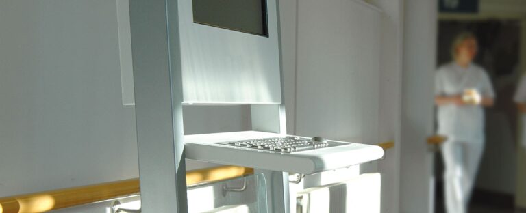Väggmonterad datorpulpet i metall med inbyggt tangentbord i sjukhusmiljö, med vårdpersonal synlig i bakgrunden.